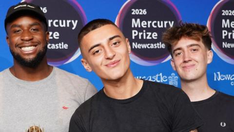 Knats standing in front of a blue 2026 Mercury Prize board and smiling. King David-Ike Elechi is on the left and has a black, short beard and is wearing a black cap. He is wearing a grey jumper and is holding the gold Mercury Prize nomination trophy. Stan Woodward is standing in the middle and is wearing a black t-shirt and has tattoos on both arms. Ferg Kilsbyis on the right and is wearing a black t-shirt.