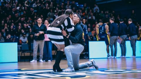 Man is down on one knee proposing to his partner in a basketball arena