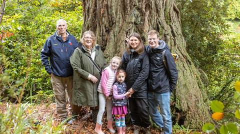 Four adults and two children stand in front of a redwood tree in the middle of a garden surrounded by other trees and plants.
L-R: James Weatherhead (son), Elaine Weatherhead (daughter,-in-law), Maddie Hollis, Eliza Hollis (great granddaughters), Amy Hollis (granddaughter) and Jack Hollis (grandson- in-law)
