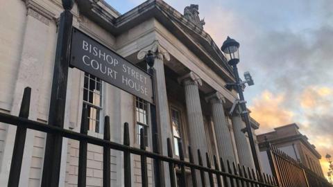 A sign for Bishop Street courthouse looms over black iron railings. Behind the railing the front of Derry's courthouse can be seen, with a lampost in front of four large pillars.