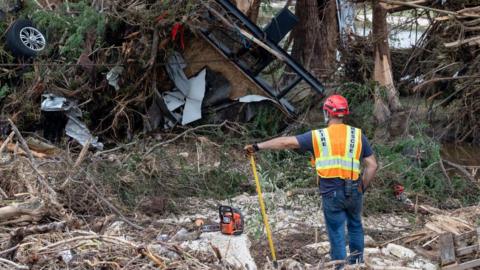 A rescuer worker in orange high vis stands in front of wreckage in a forested area, towering far above him
