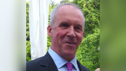 John Murkin is smiling at the camera while wearing a blue blazer, purple tie and light blue shirt. He has short grey hair and is standing outside in front of green trees and bushes. 