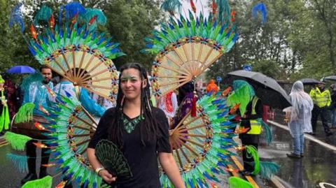 A woman wearing bright green and blue feathered wings which radiate in circles from her and shoulders. She is also carrying a fan and her face is decorated with jewels and face-paint. Her long black hair is braided and she is walking down the centre of a rainy street, with others processing behind her and onlookers lining the pavements.