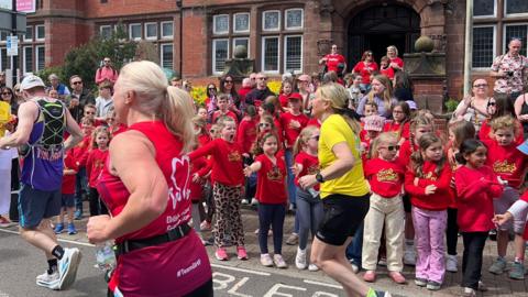 A group of dozens of little girls in red tops watch runners go by. Some put their hands out to high-five runners in front of an old redbrick building in Altrincham.