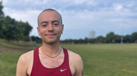 A man in a claret Nike running vest stood in a park. He has a shaved head and is wearing a chain around his neck.