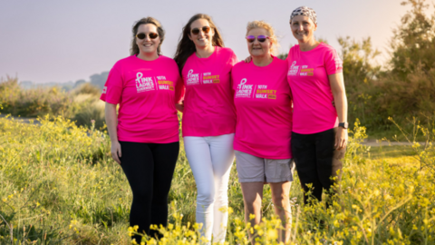 A group of four women stand in a field wearing pink The Pink Ladies sunset walk tops. The women are standing in a row.