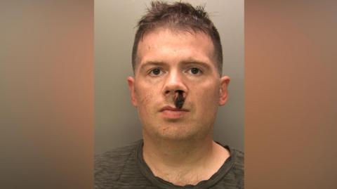 A mugshot of Patrick Young who is looking into the camera with a straight face. He has brown, short hair which is sticking up on the top. There is thick, dried blood around under his nostrils. He is wearing a grey t-shirt.