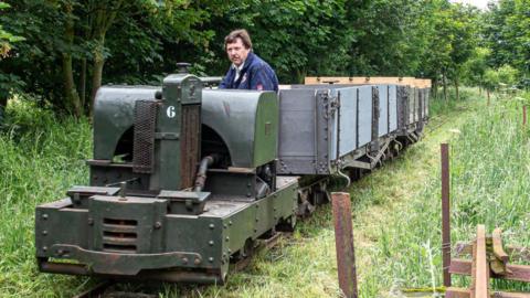 A man sat in a Simplex diesel train with two Class D bogie wagons being carried behind on a train track, surrounded by grass and trees