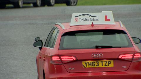 A red car with a sign on top which says 'NI Driving School' and an L sign