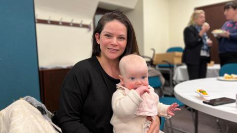 Cerin smiles at the camera as she sits by a white table with her seven month old baby Clementine on her lap. Cerin is wearing a black jumper and has long brown hair. Clementine is wearing a white baby jumper and has a light pink bib on.