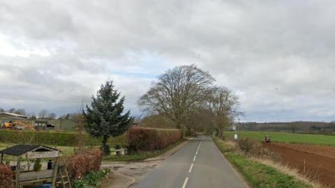 A country road with a farm on one side and fields and trees on either side. 