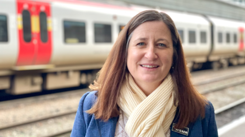 A woman with long brown hair, a cream scarf and blue coat on a railway platform with the blurred shape of a grey train behind her