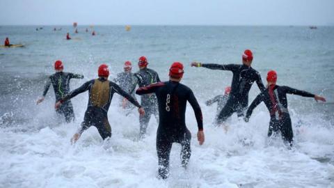 A group of participants wading into the sea wearing wetsuits and red swimming caps as they compete in the swim leg of the race of the IRONMAN 70.3 Weymouth in 2018. People can be seen further out to sea in the distance. 