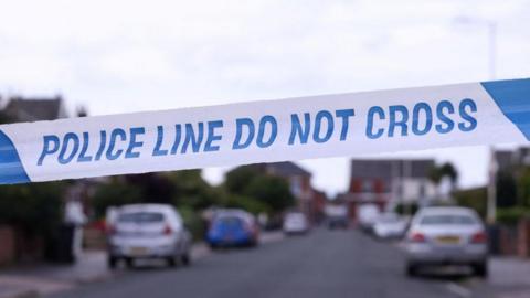 Police tape across a road. It reads, police line do not cross. Cars can be seen parked on the street behind the tape.