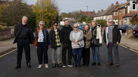 Eight residents stand in a row in the road with houses lining the side of the street. There are a mixture of men and women. The road features single yellow lines. 