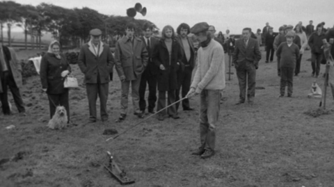 A black-and-white still from the TV programme Nationwide in 1972. A crowd of people are watching a man in a flat cap with a stick attempt to hit a small ball-like object off a spring-based wooden trap.