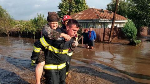 A Romanian rescuer carries an elderly woman on his back out of a flooded street, while a couple of neighbours look on in front of their house