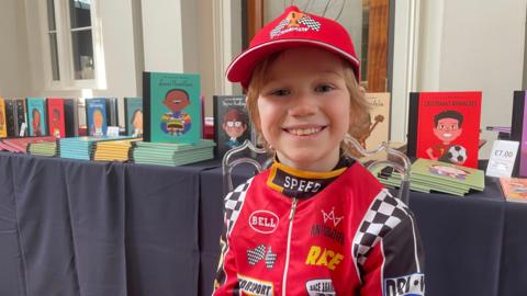 A young child in a red racing driver costume and cap grins at the camera, standing in front of a table displaying Little People, BIG DREAMS books including titles on Lewis Hamilton, Stephen Hawking, Nelson Mandela and Cristiano Ronaldo.
