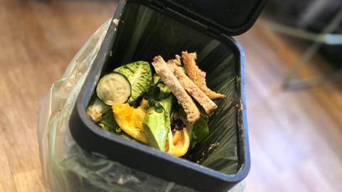 Food waste in an open bin, including bread, courgette slices and salad leaves. There is a wooden floor and the food is contained within a bag inside the bin.