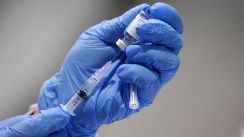 A blue disposable glove holding a vaccination needle and syringe that is being filled with a clear liquid. 