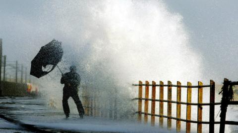 A man dressed all in black struggles to control a black umbrella next to a huge wave coming over a sea wall.