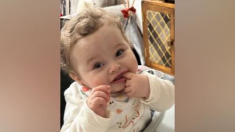 Baby with curly hair sitting in high chair - he has his finger in his mouth. He is wearing a baby grow with an elephant on it.