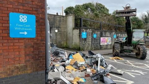 A telehandler is seen next to a damaged brick wall. There is a sign that says "free cash withdrawals".
