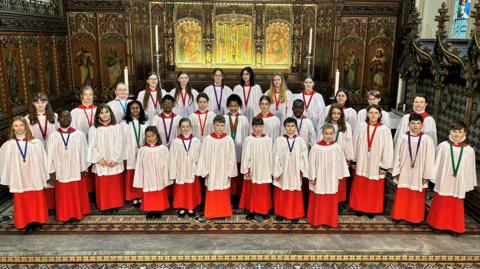 A group of young choristers wearing red and white robes. They smile at the camera as they stand within a church.