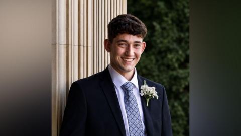 A man with short brown hair smiles at the camera. He is wearing a suit with a blue tie and appears to be standing outside.