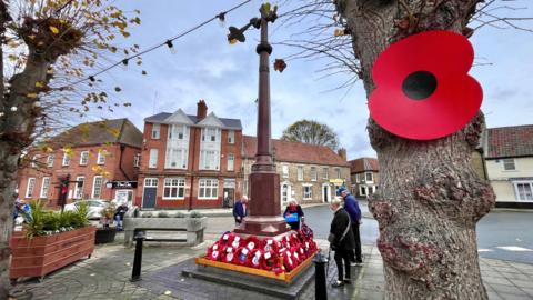 A large red poppy is pinned to a tree in the foreground. In the background a small group of people gather around a tall war memorial in a town centre. It has lots of poppy wreaths at its base.