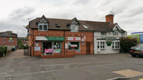 A building in the middle of the photo has the words post office in white on a green backing and to the right of that is a yellow and red post office logo.