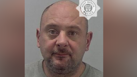 A mugshot of a man with very short hair and a stubbly beard. He is wearing a grey prison jumper and is against a neutral grey background. 