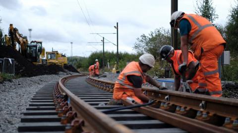 Five engineers in orange high-vis work on railway track with yellow diggers in the distance.