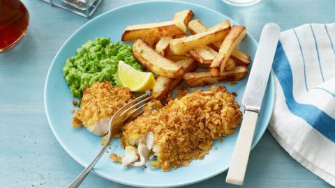 A plate of fish and chips with mushy peas and a wedge of lemon. There is cutlery and a tea towel beside it.