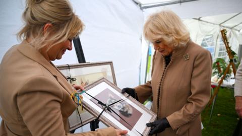 Camilla wears a brown jacket with a glittery broach and holds a portrait of a small dog. Another woman in a brown jacket also holds the portrait.
