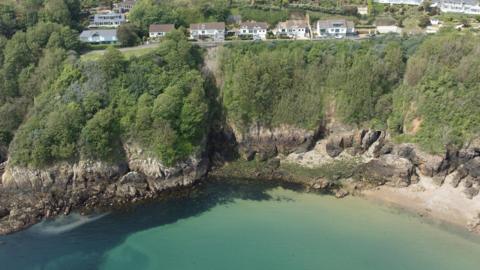 Soldier's Bay and the cliff face above it. At the top is Fort George a collection of large houses with brown roofs and white facades. Beneath the houses is a layer of greenery, with trees covering the middle half of the cliffs, above brown strips of granite getting darker as it goes closer to an azure blue sea. 