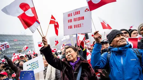 A woman in a brown fur coat waves a Greenlandic flag during a protest in Nuuk. There's a large crowd around her, also waving the red and white flag and holding placards