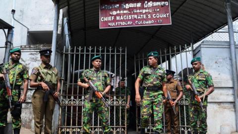 Men dressed in camouflage uniform holding rifles in front of metal gates of a court