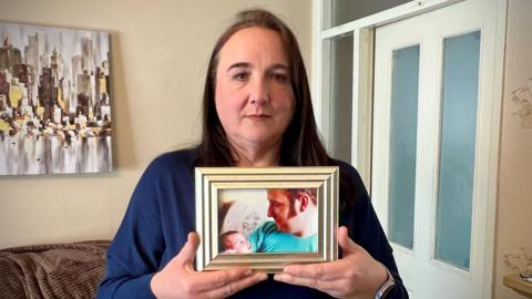 A woman in her fifties is standing in a living room holding up a picture of a man cuddling a baby. The man in the picture is her dad and she is the child. Behind her is a door to the kitchen and there is a sofa pushed up against a wall.