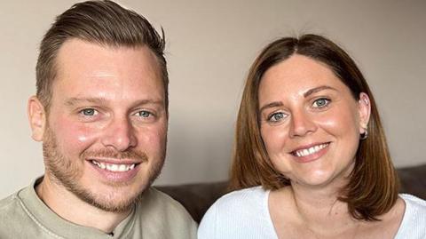 Ben Uttley and his younger sister Holly Murdy, sitting on a brown sofa and smiling. Behind them is an out-of-focus white cushion covered with a design of small black triangles. He has short, dark blonde hair, with the front combed back, and has a moustache and short beard. He's wearing a pale khaki t-shirt. She has shoulder length light brown hair in a side parting and is wearing a white, v-neck button up top.