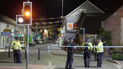 Police in yellow high vis coats standing on a road behind police tape.