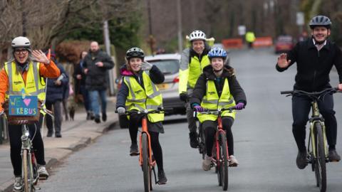 A woman, two men and two girls wear high vis vests and helmets and are riding their bicycles along a road. They are smiling and waving. 