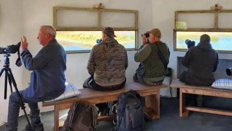 Four men sitting, birdwatching, through windows in the hide; one man has his camera on a tripod. The hide overlooks a pond and grassland.