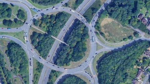 The road junction seen from the air, with a roundabout busy with traffic passing over a trunk road. There are five exits off the roundabout, two of which head to residential areas visible to the right. The land in between the roads is covered in trees and shrubs.