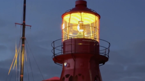 The top of a lighthouse is lit up at dusk. It's glowing with an orange light and a ship's mast can be seen to the left of it. The sky is cloudy and a dark blue-grey colour.