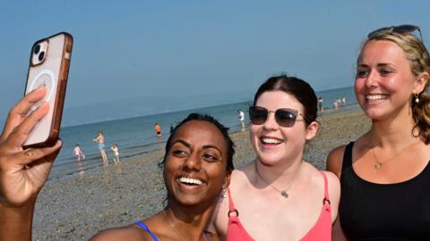 Three girls on a beach taking a selfie. They are all in swimsuits. Two have dark hair and one has blond hair. They are all smiling for the camera.