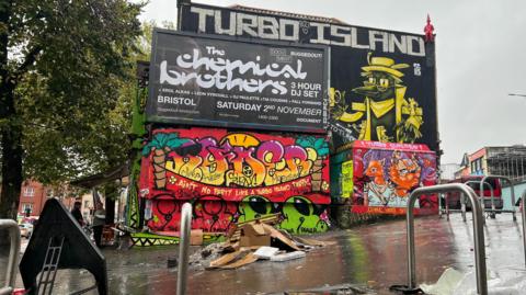 Graffiti and an advertising sign on a wall in Bristol, with rubbish spewed on the floor.