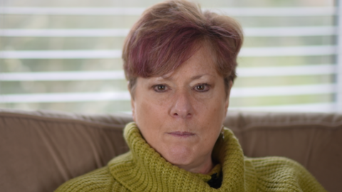Lynne O'Neill has short hair and looks directly into the camera. She is wearing a green roll-neck sweater and sitting on a brown sofa with a white Venetian blind behind her.