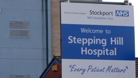 A view of a blue and white sign for Stepping Hill Hospital in Stockport with the words 'every patient matters'. The sign is set in front of a red brick outbuilding and a larger hospital building. 