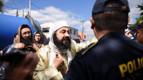 A member of the Lev Tahor sect, bearded and wearing a yellow robe and white headscarf, remonstrates with a Guatemalan police officer dressed in a blue uniform with his back to the camera